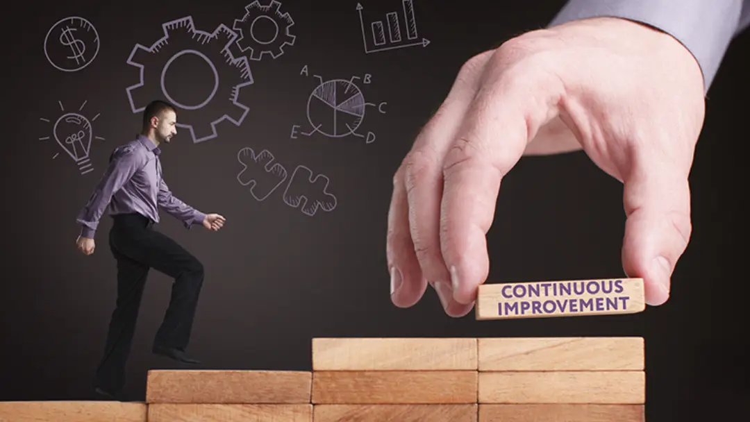 Studio image of a person walking up on some wooden blocks with a large hand placing a block reading continuous improvement.