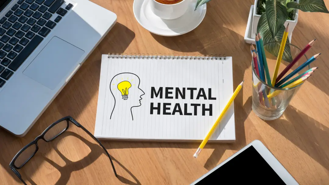 The studio captures a view of a desk adorned with a notepad, showcasing an outline of a person and the phrase "mental health" in a doodle-style font. There are coloured pencils in a jar next to the pad.