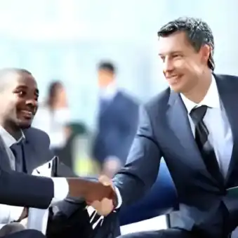 The image captures a close-up of office workers sitting in a casual meeting; one person is shaking hands with one other person. There are other persons in the background.